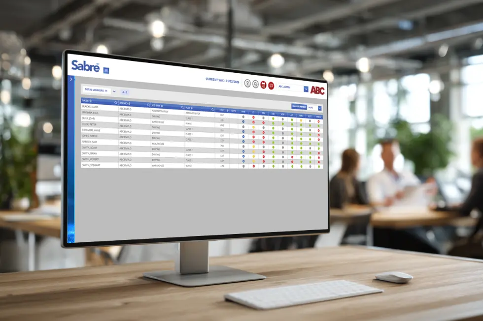 A modern office setting showing the Sabre MS recruitment software dashboard on a computer screen, used for managing temporary staffing bookings and compliance